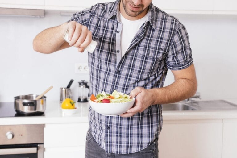 Seorang pria berkemeja kotak kotak menaburkan bumbu ke semangkuk salad di dapur modern, menyadari bahwa diet tinggi garam dapat meningkatkan risiko batu ginjal.