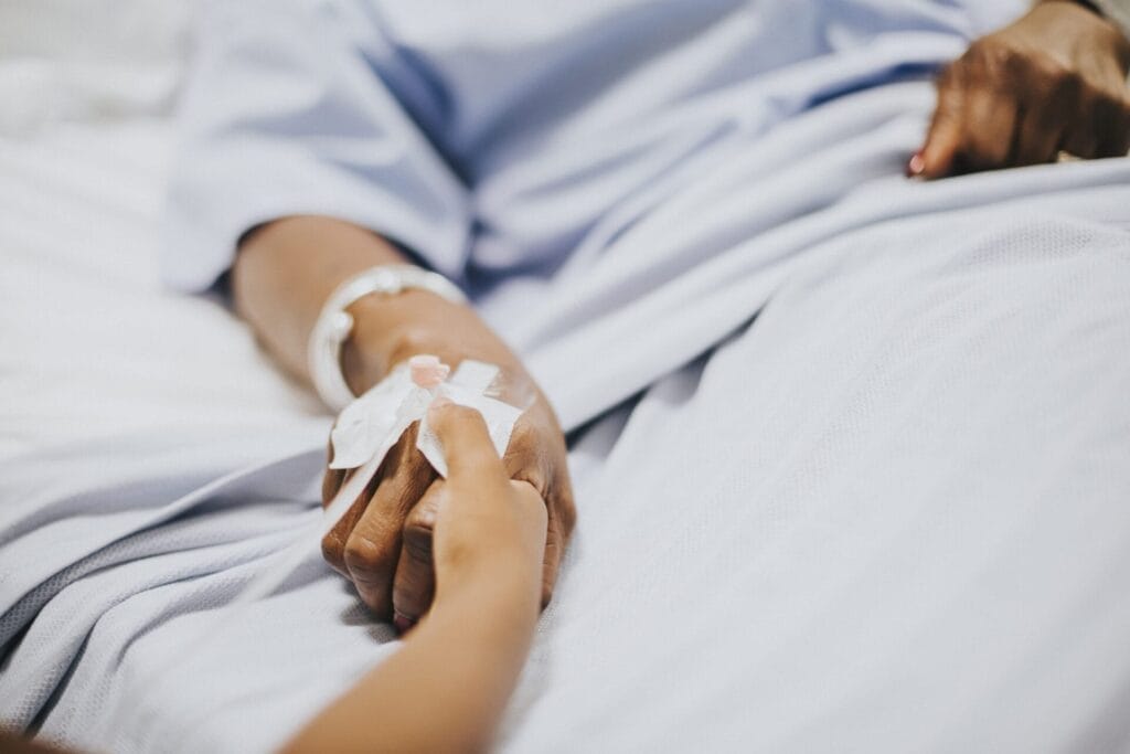 Seseorang yang mengenakan gaun rumah sakit dengan infus di tangannya, mungkin menghadapi risiko sepsis atau komplikasi batu ginjal, memegang tangan orang lain sambil berbaring di ranjang rumah sakit.