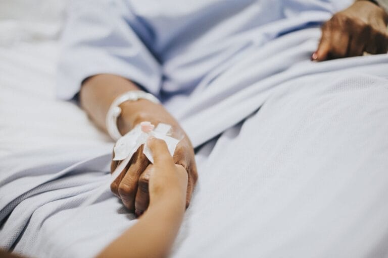 Seseorang yang mengenakan gaun rumah sakit dengan infus di tangannya, mungkin menghadapi risiko sepsis atau komplikasi batu ginjal, memegang tangan orang lain sambil berbaring di ranjang rumah sakit.