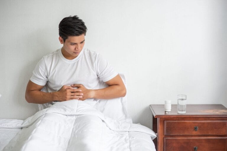 Seorang pria duduk di tempat tidur sambil memegang perutnya dengan kedua tangan, tampak tidak nyaman—mungkin karena batu ginjal. segelas air dan sebotol obat ada di meja samping tempat tidur di sampingnya.