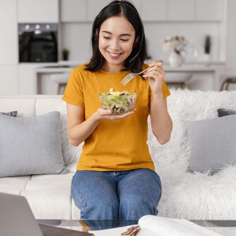 Seorang wanita mengenakan headset duduk di sofa, tersenyum saat dia memakan salad dari mangkuk—mungkin mengikuti diet untuk mencegah batu ginjal—dengan laptop dan dokumen di atas meja di depannya.