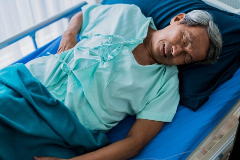 Lansia terbaring di kasur rumah sakit dengan mata tertutup, mengenakan baju pasien dan selimut biru, menunjukkan gejala infeksi saluran kemih.