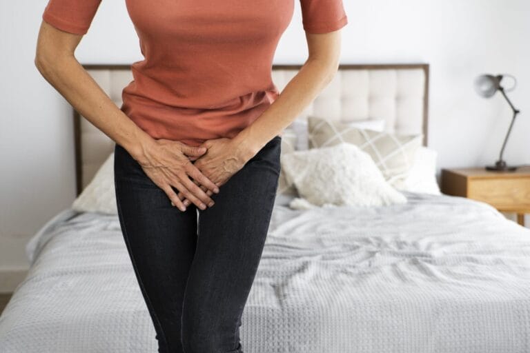 Seorang wanita berdiri di depan tempat tidur, memegang perut bagian bawahnya dengan kedua tangan, menunjukkan rasa tidak nyaman atau nyeri akibat infeksi saluran kemih.