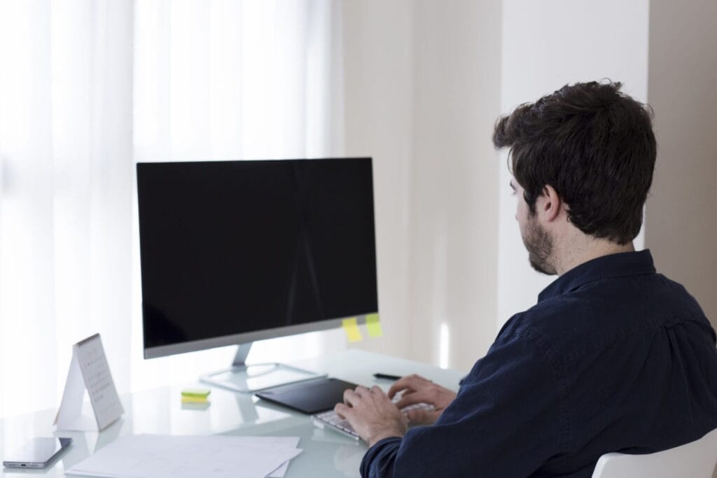 Gambar saat ini: Seorang pria berambut hitam duduk di meja mengetik pada keyboard di depan monitor komputer yang dimatikan di kantor modern yang terang, menggambarkan risiko gaya hidup sedentari yang dapat meningkatkan risiko terkena batu ginjal.