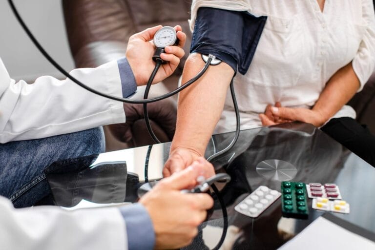 Seorang dokter mengukur tekanan darah pasien dengan alat tensimeter di klinik, di atas meja terlihat beberapa jenis obat.