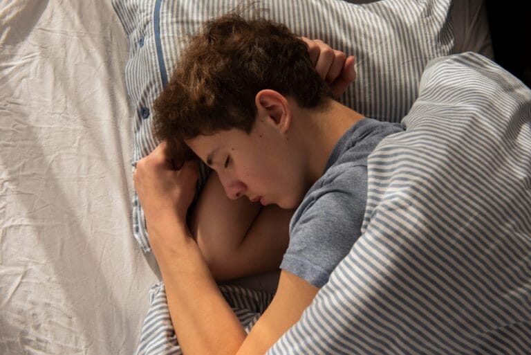 Remaja laki-laki tertidur di tempat tidur dengan posisi meringkuk, menggambarkan kondisi enuresis atau mengompol saat tidur malam