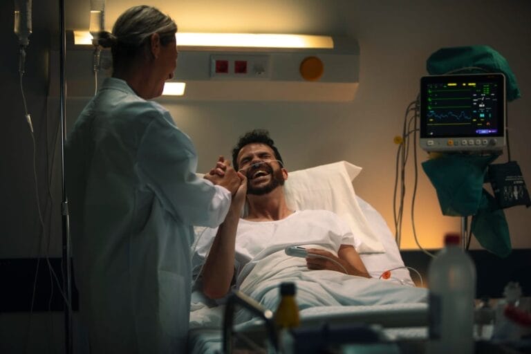 Pasien pria sedang tersenyum di ranjang rumah sakit sambil menjabat tangan dokter, dengan alat monitor medis dan infus di sekitarnya.