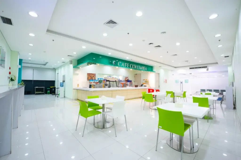 Kafe modern yang terang benderang dengan meja putih, kursi hijau dan putih, serta konter berlabel "Café Columbia" di latar belakangnya. Tempat yang menarik ini juga dikenal sebagai tempat untuk berdiskusi tentang Penanganan Batu Ginjal dalam suasana yang bersih dan luas.