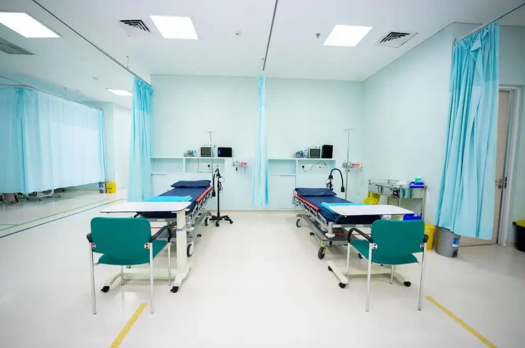 Kamar rumah sakit yang bersih dan terang benderang dengan dua tempat tidur pasien kosong yang dipisahkan oleh tirai biru, peralatan medis untuk penanganan batu ginjal, dan kursi berwarna teal.