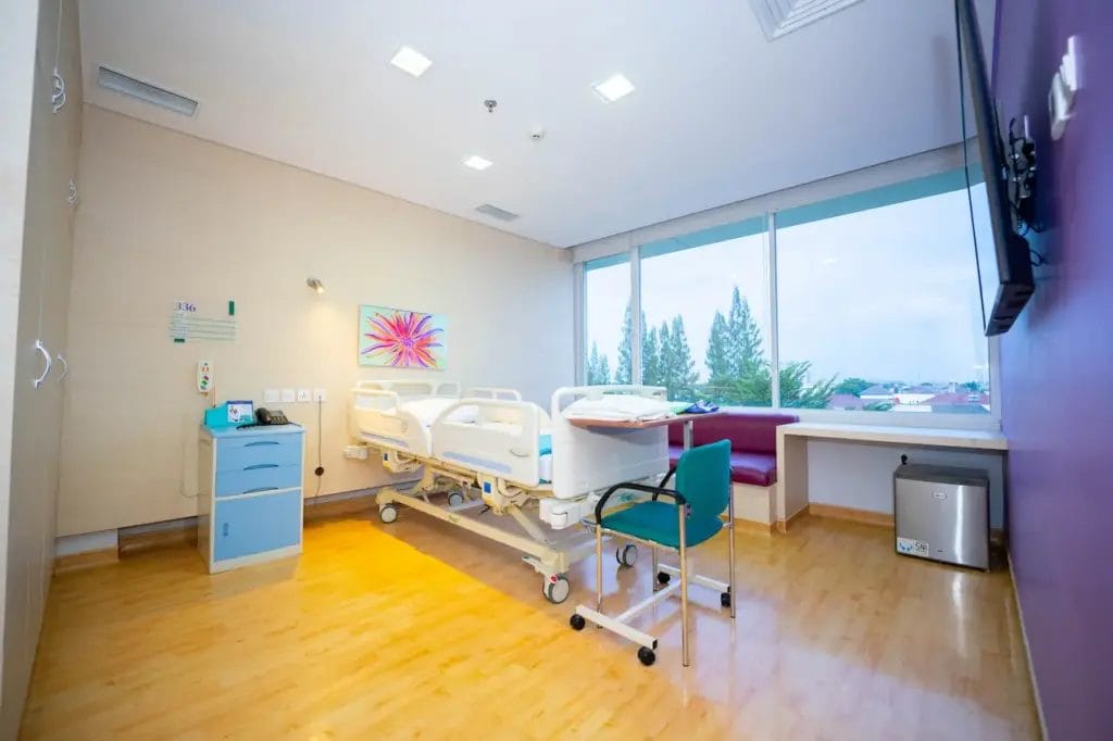 Kamar rumah sakit yang dirancang untuk Penanganan Batu Ginjal dilengkapi dengan tempat tidur tunggal, peralatan medis, kursi hijau, meja samping, TV yang terpasang di dinding, jendela besar, dan lantai kayu.