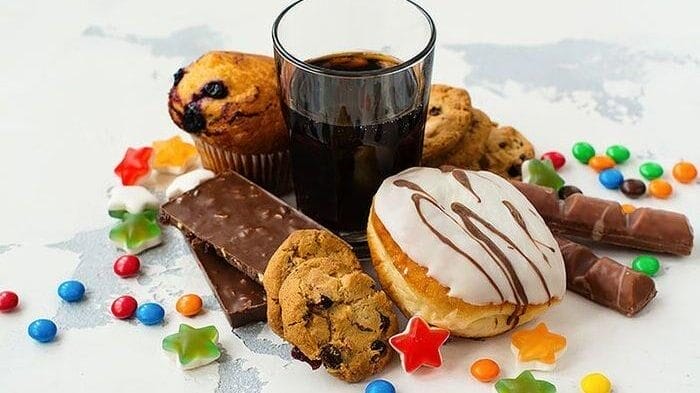 Segelas soda hitam yang dikelilingi oleh kue kering, donat, muffin, cokelat batangan, dan permen warna warni di atas permukaan putih, secara otomatis menangkap konsep kenikmatan camilan yang mudah.