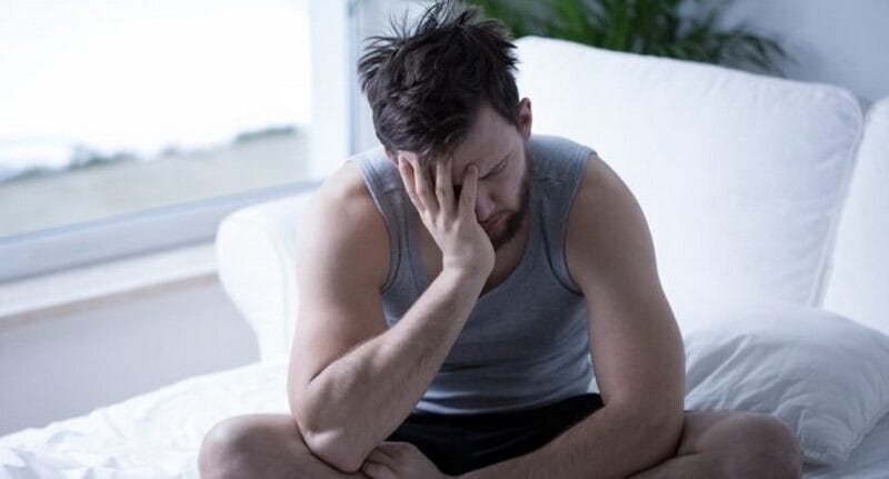 Seorang pria dengan tank top abu abu duduk di atas tempat tidur dengan tangan menutupi wajahnya, tampak lelah atau stres, seolah olah terbebani oleh tekanan Konsep Otomatis dalam rutinitas hariannya.