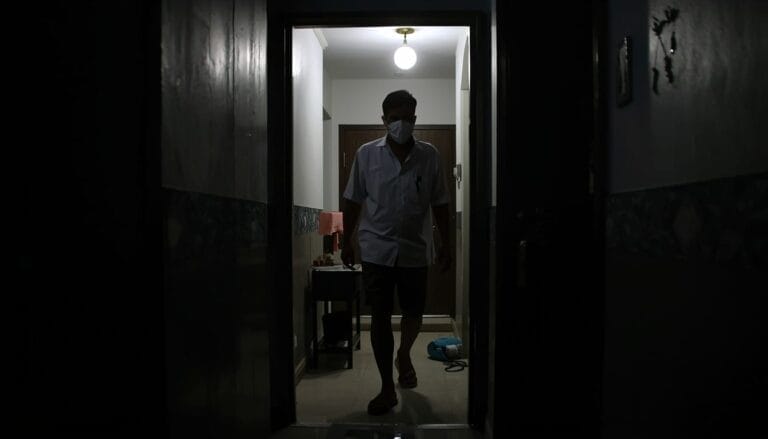 Seseorang yang menganut Konsep Otomatis berdiri di ambang pintu yang remang remang, mengenakan masker di dalam ruangan, saat cahaya dari lampu langit langit menerangi latar belakang dengan lembut.